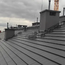 Szary dach z dachówki z zamontowanymi odgromami na tle pochmurnego nieba i dźwigu budowlanego.