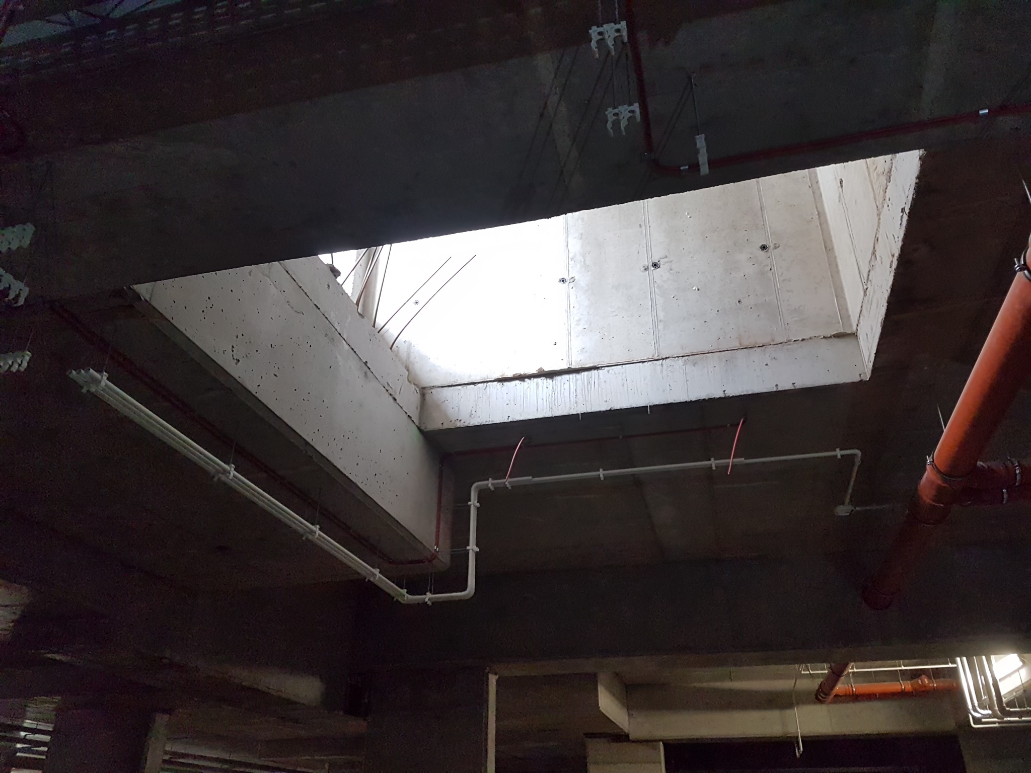 Betonowy sufit z widocznymi instalacjami: rurami, przewodami w białych listwach oraz otwartym szybem wentylacyjnym w górnej części kadru.