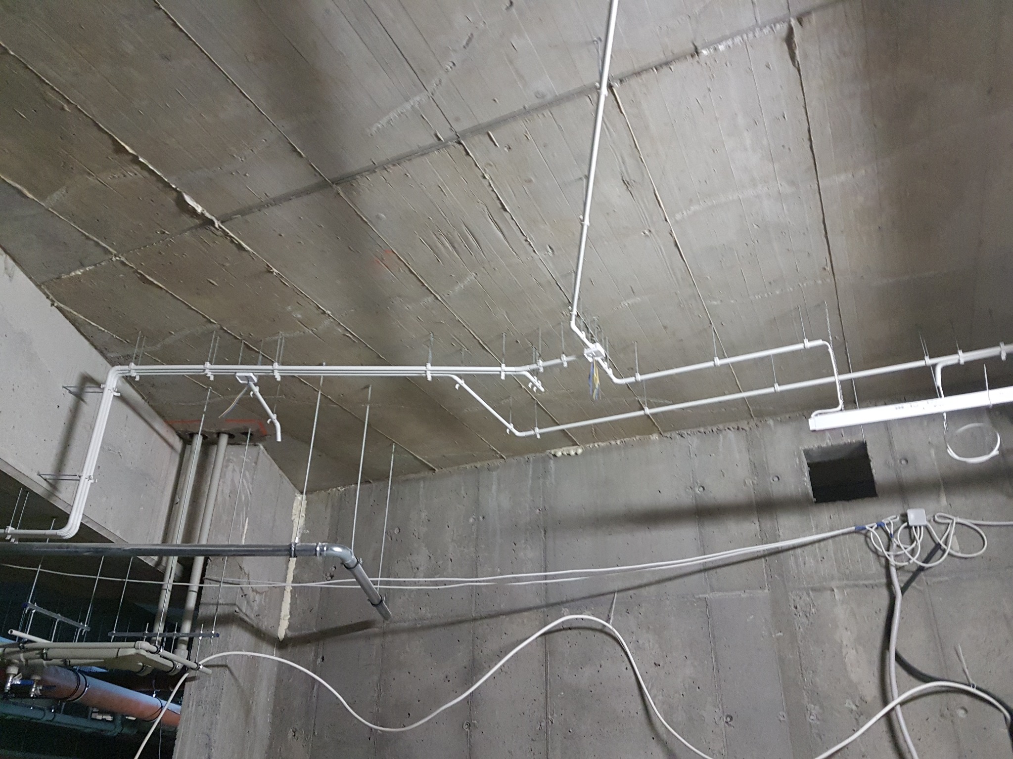 Surowy betonowy sufit i ściany z widocznymi rurami i przewodami elektrycznymi prowadzonymi natynkowo, w trakcie instalacji.