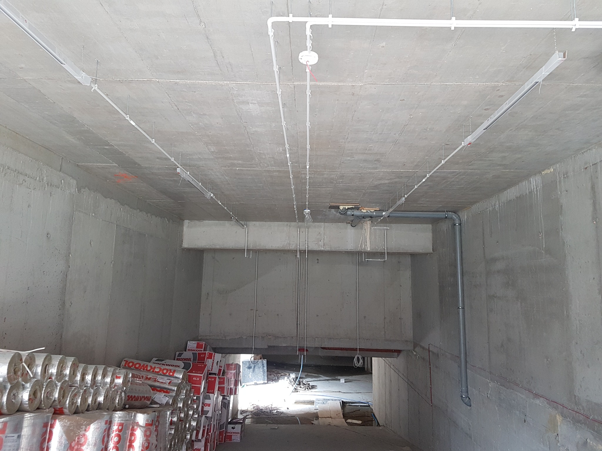 Surowy betonowy tunel z instalacją elektryczną pod sufitem, widoczne kable, rury i rolki izolacji Rockwool