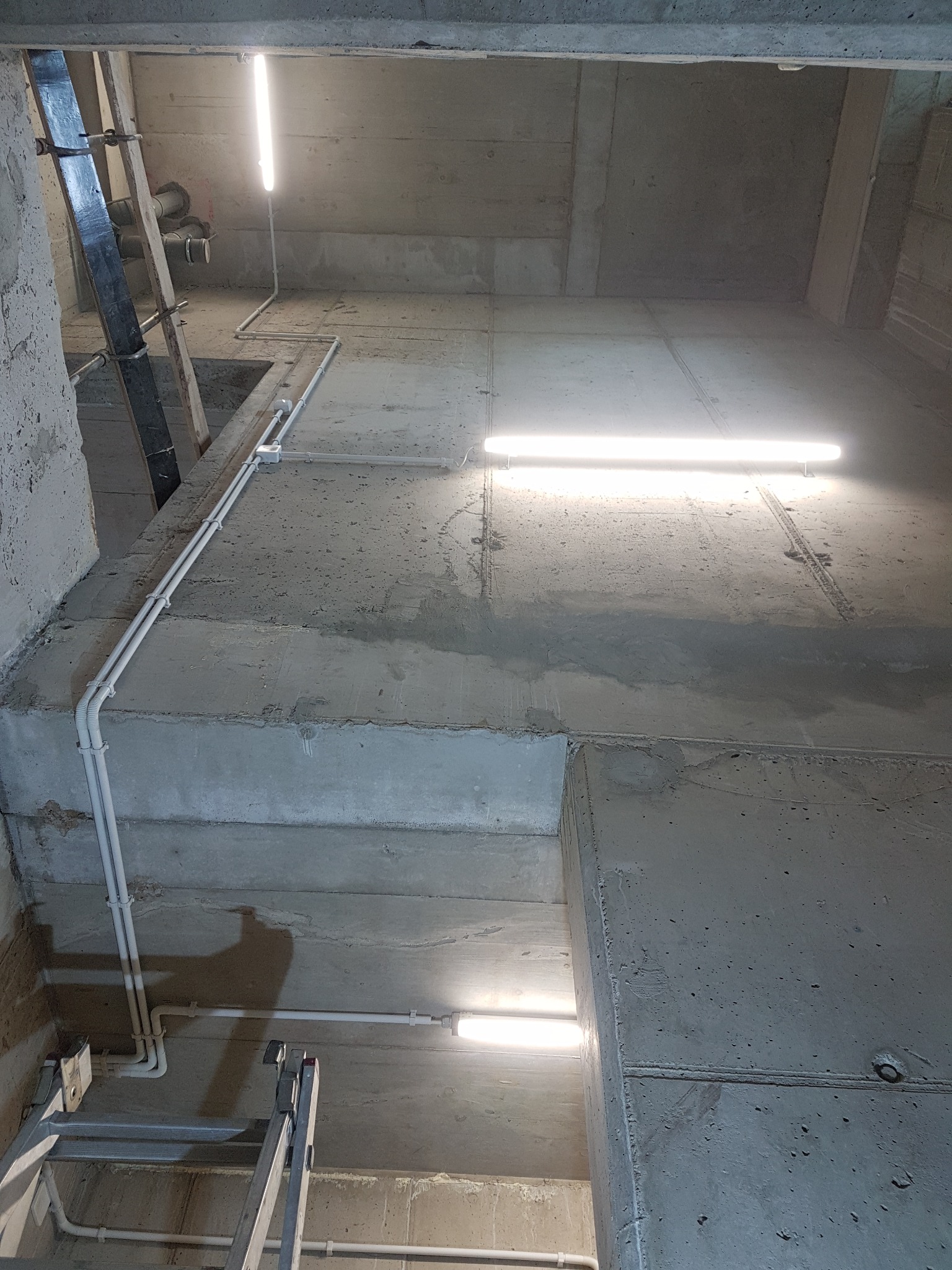 Surowy betonowy strop z widocznymi, prowadzonymi po powierzchni przewodami elektrycznymi w białych rurkach PCV, oświetlony podłużnymi lampami LED. Widoczna drabina oparta o ścianę.