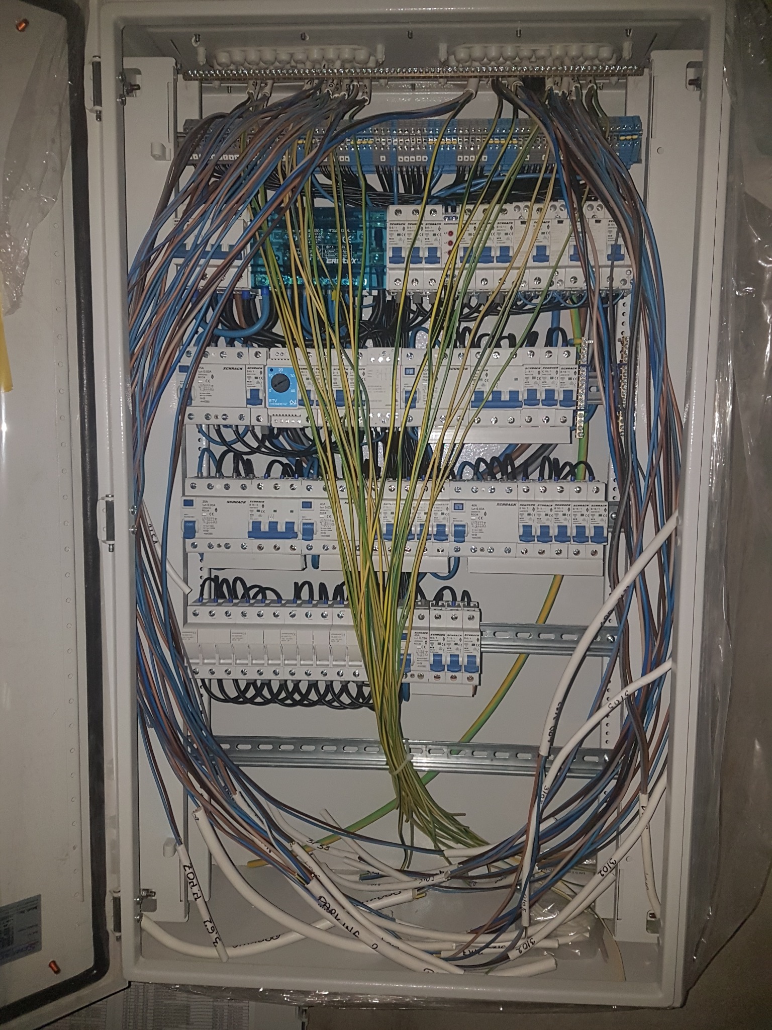 Otwarta skrzynka elektryczna z licznymi przewodami w kolorach niebieskim, brązowym, żółto-zielonym i białym, częściowo uporządkowanymi w wiązki, z widocznymi bezpiecznikami i elementami instalacji...
