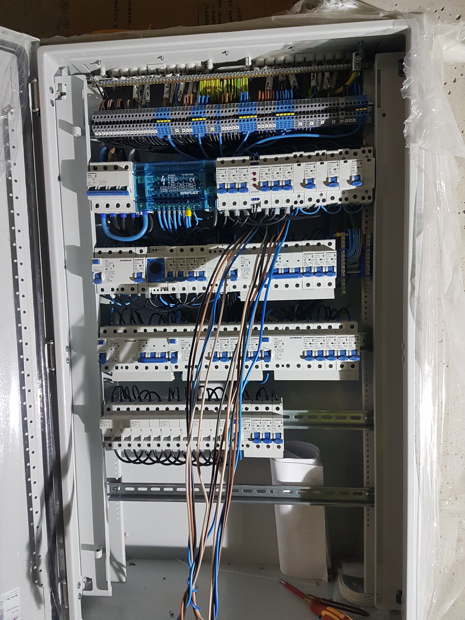 Otwarta skrzynka elektryczna z licznymi bezpiecznikami, przewodami i listwami zaciskowymi w Świebodzinie. Część przewodów zwisa luźno przed skrzynką. Widoczny biały pojemnik i czerwono-żółty...