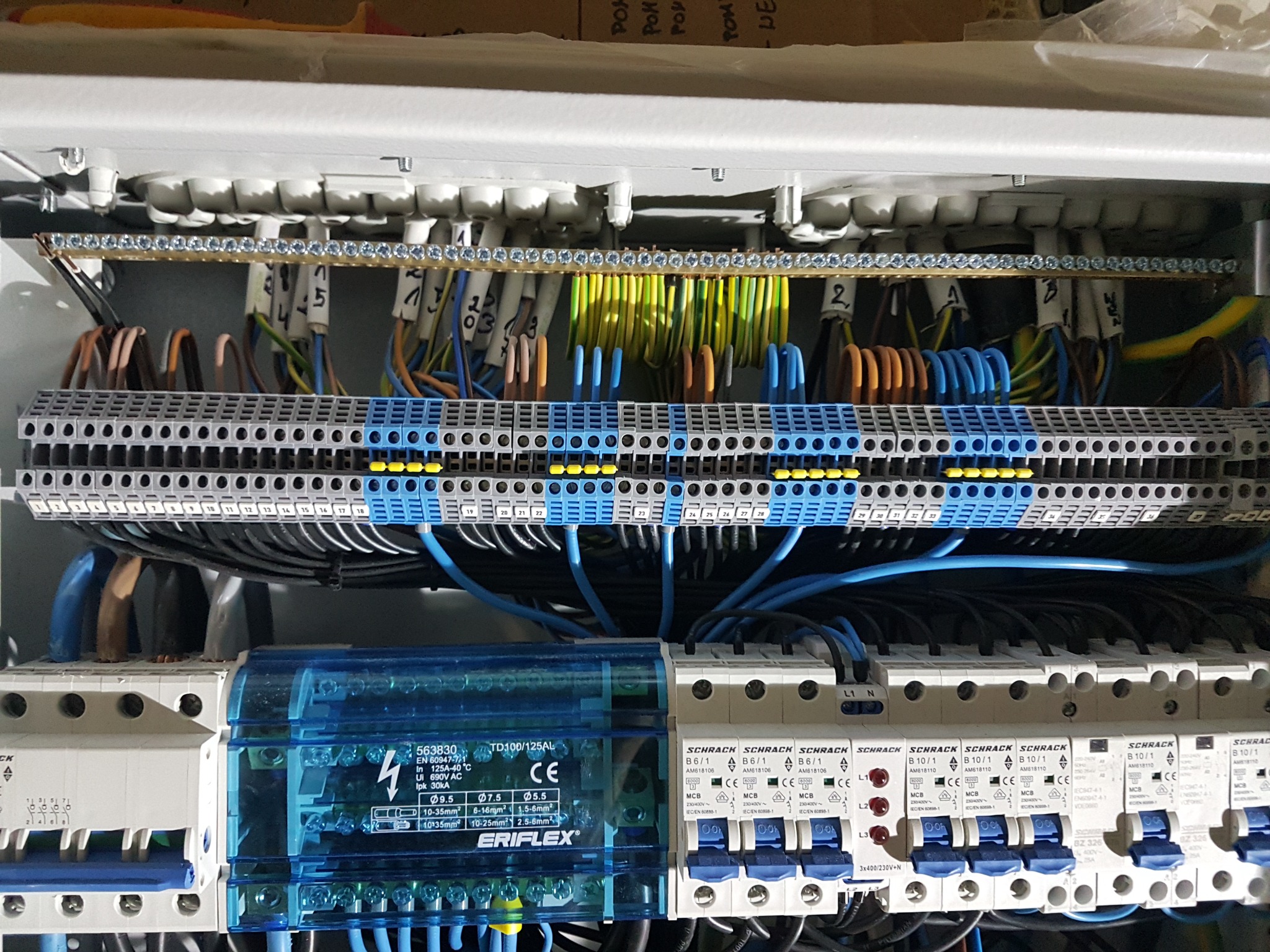 Rozdzielnia elektryczna z widocznymi bezpiecznikami, listwami zaciskowymi i okablowaniem w kolorach niebieskim, żółto-zielonym i brązowym, z etykietami na przewodach.