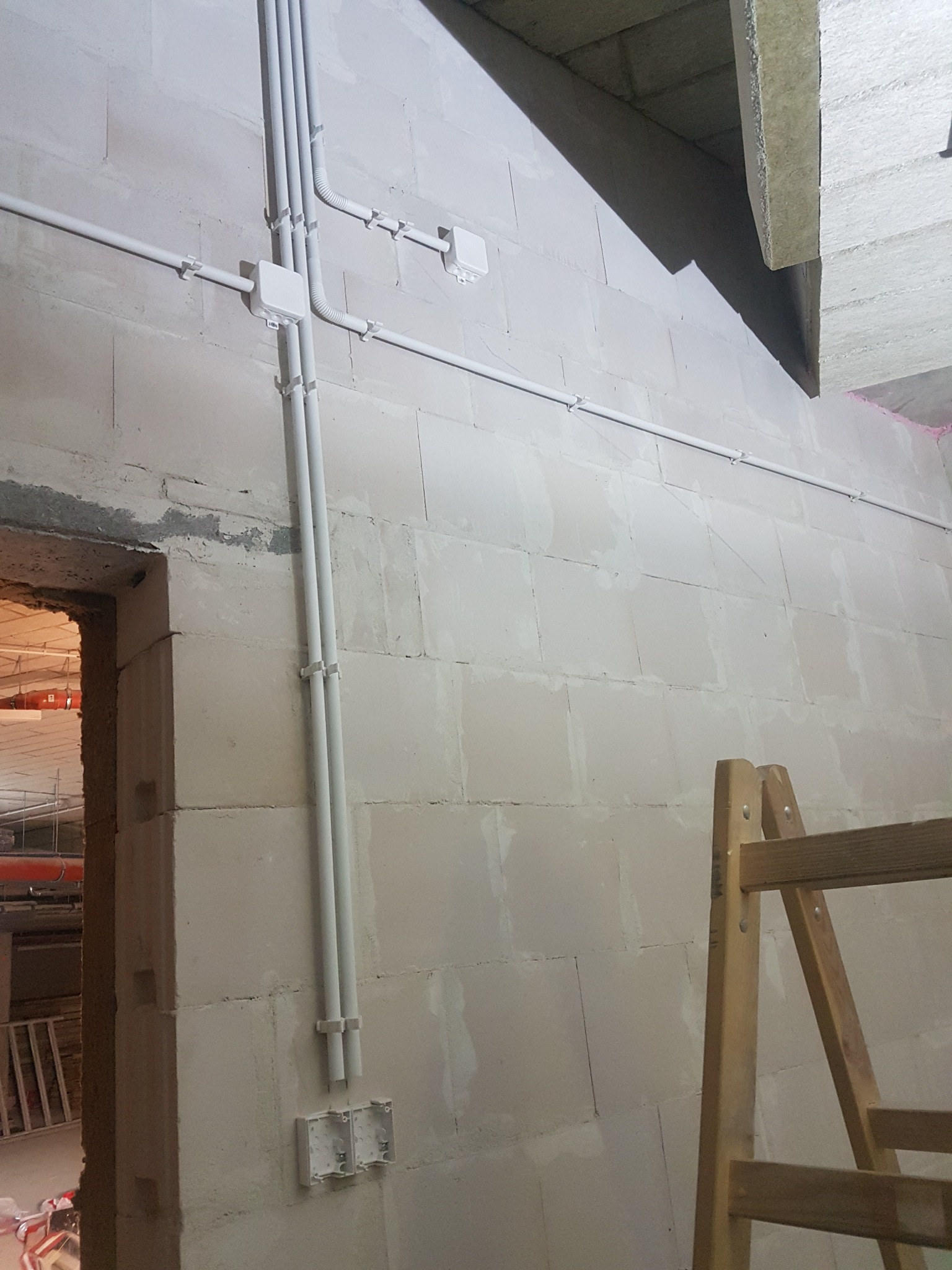 Surowe ściany z betonu komórkowego z widoczną instalacją elektryczną w białych rurkach PCV prowadzonych natynkowo, puszki elektryczne bez osprzętu, fragment drabiny drewnianej.