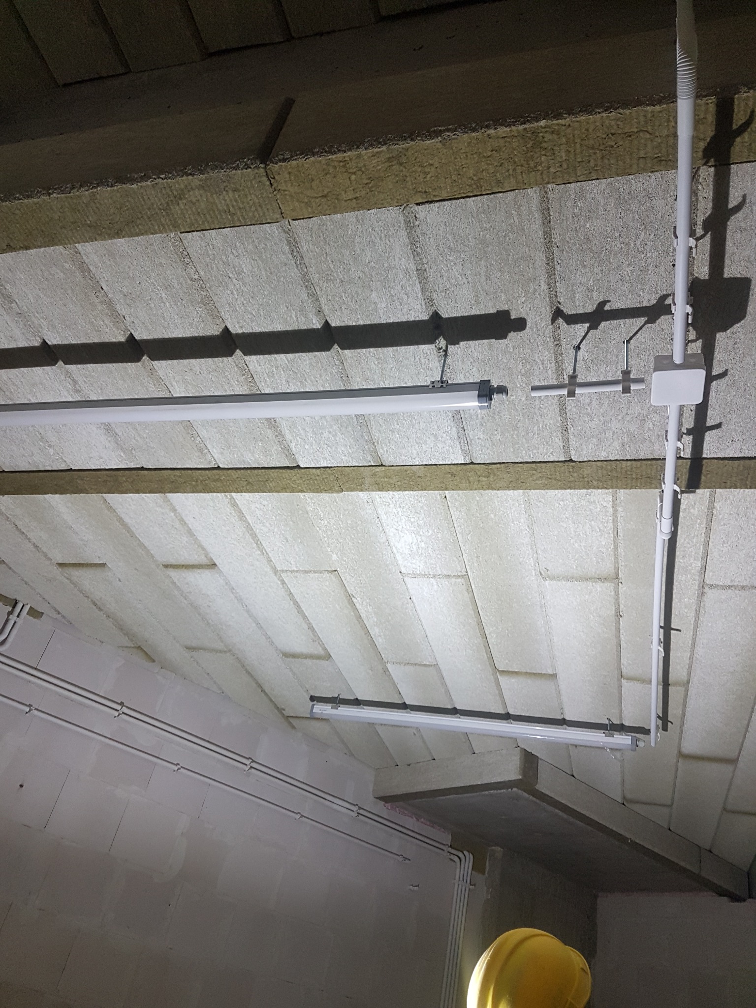 Montaż oświetlenia w pomieszczeniu z izolacją stropu, widoczne rury instalacyjne i fragment kasku ochronnego.