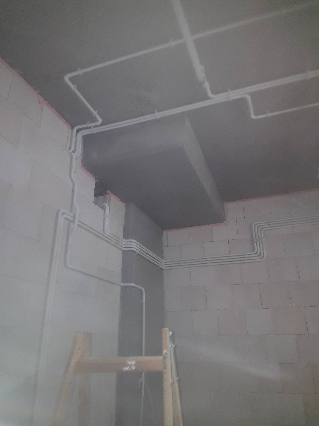 Białe rurki instalacji elektrycznej ułożone na surowym, szarym suficie i ścianach z pustaków, widoczna drabina drewniana.