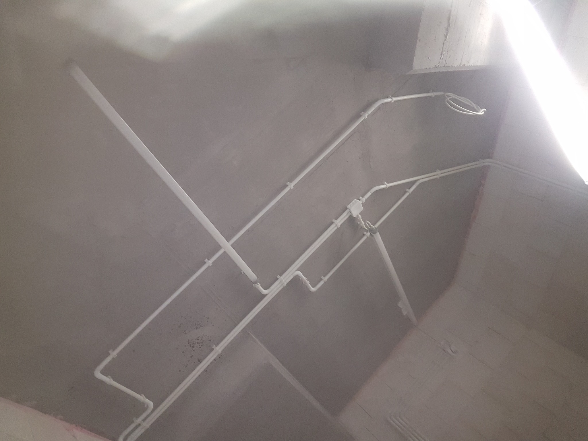 Białe rury instalacji elektrycznej przymocowane do surowego, szarego sufitu w nowym budynku, widoczne łączenia i uchwyty mocujące.