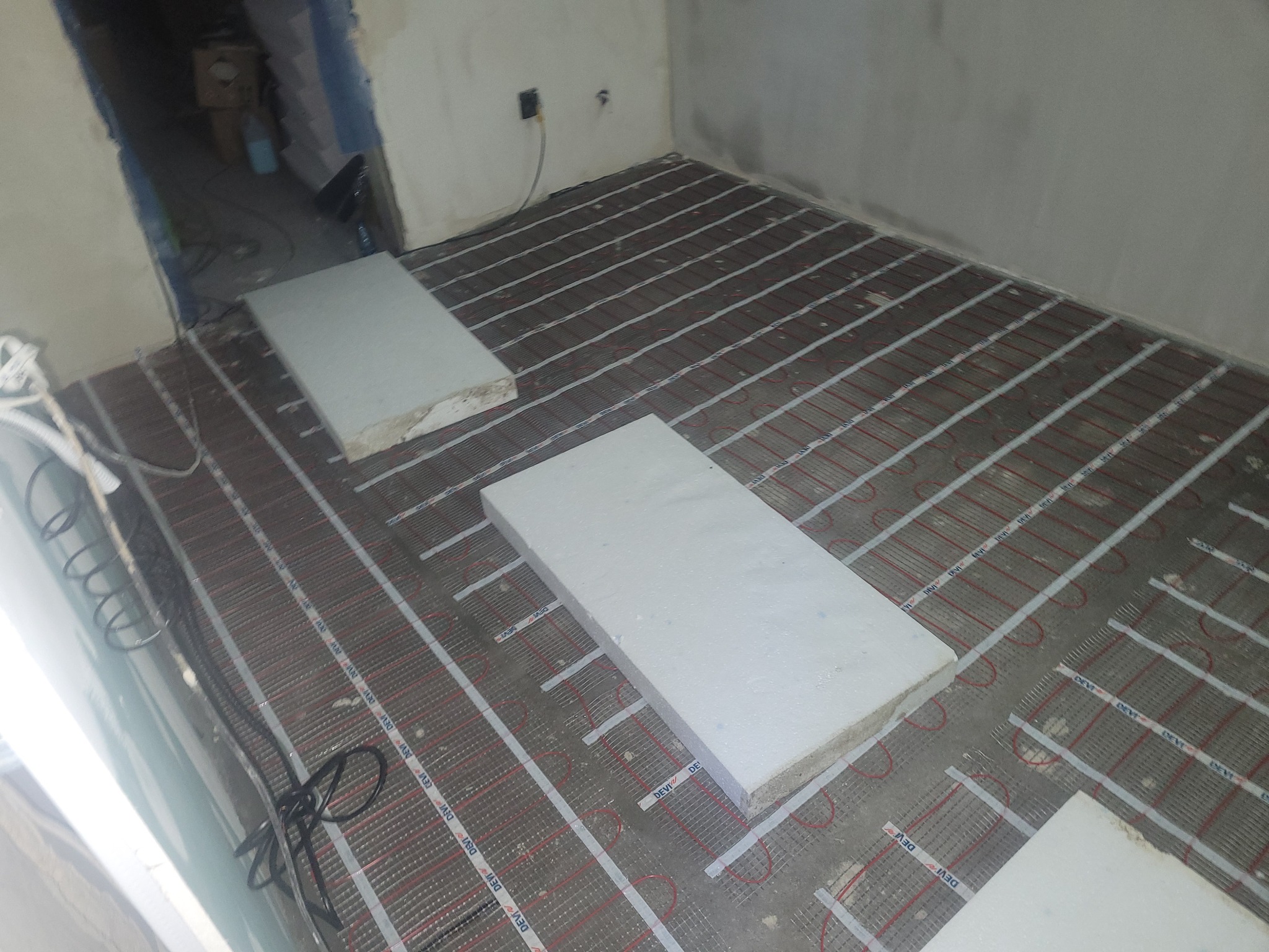 Rozłożona mata grzewcza DEVI na podłodze w trakcie montażu, częściowo przykryta białymi płytami izolacyjnymi, widoczne przewody elektryczne i puszka podłączeniowa na ścianie.