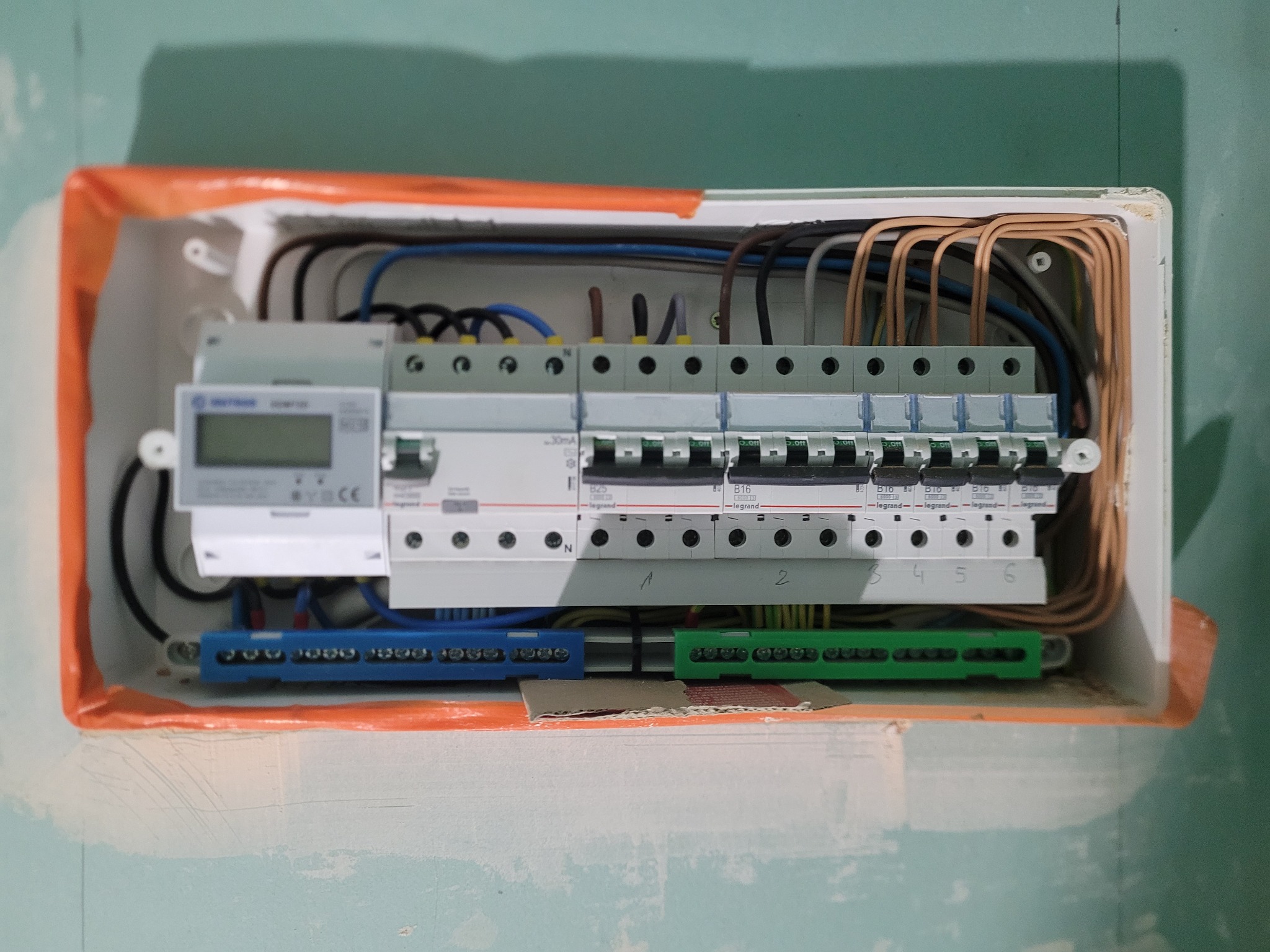 Otwarta skrzynka elektryczna z zainstalowanymi bezpiecznikami, licznikiem i okablowaniem, widoczne numerowane listwy zaciskowe i zabezpieczenia Legrand, montowana na surowej ścianie.