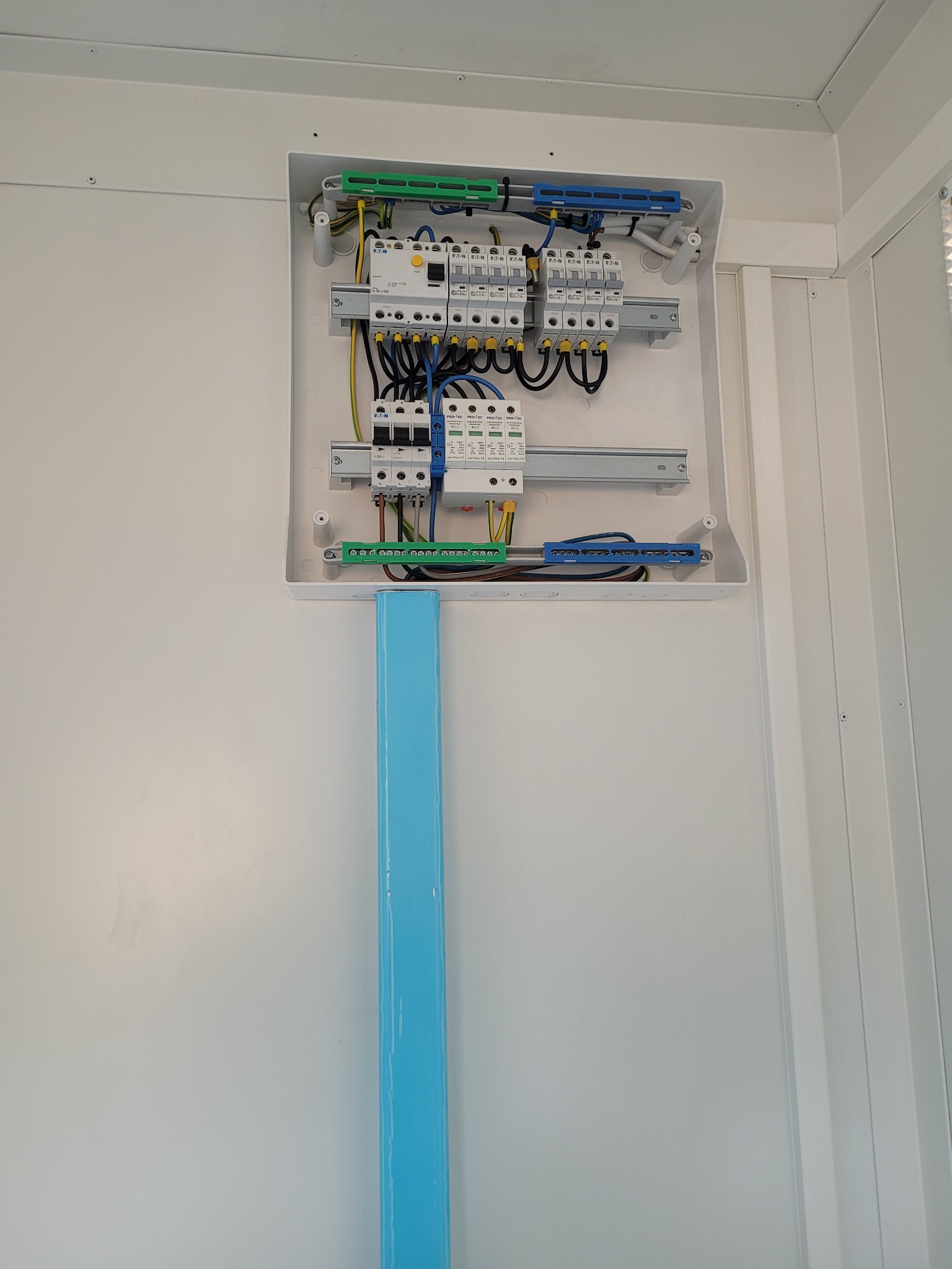 Otwarta skrzynka elektryczna z zainstalowanymi bezpiecznikami i przewodami, umieszczona na białej ścianie z niebieską rurą pod spodem.