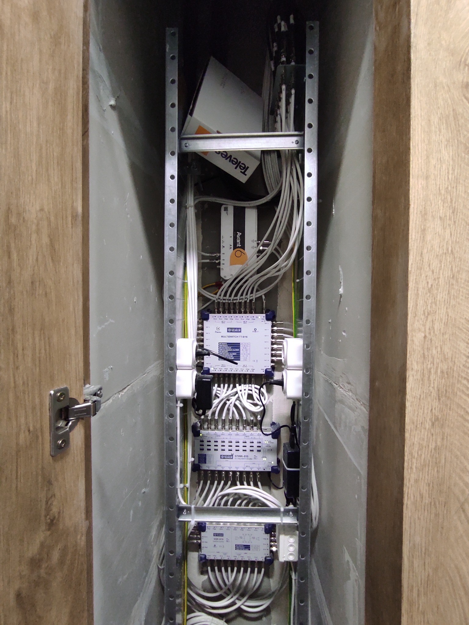Rozdzielnia antenowa w wąskiej wnęce, widoczne multiswitche, wzmacniacz antenowy Televes Avant 6, kable koncentryczne oraz metalowa konstrukcja montażowa.