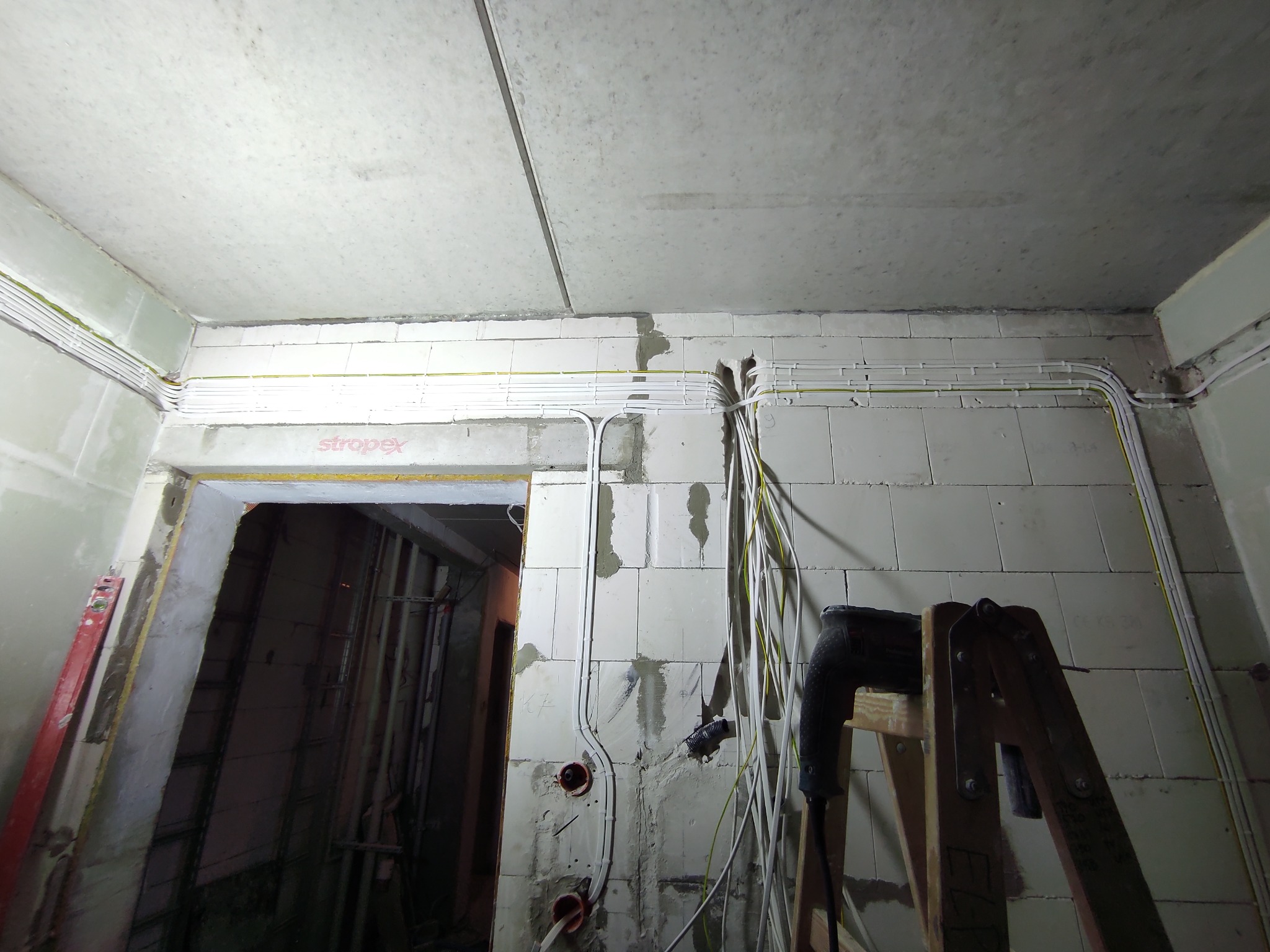 Wnętrze w trakcie remontu z widocznymi przewodami elektrycznymi w korytach i luźno wiszącymi, drabina oparta o ścianę z wiertarką, surowy betonowy strop i ściany z bloczków.