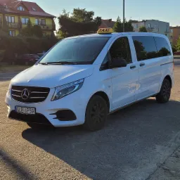 Biały van Mercedes-Benz Vito z czarnymi felgami i żółtym znakiem Taxi na dachu, zaparkowany na asfalcie w słoneczny dzień w Łebie.