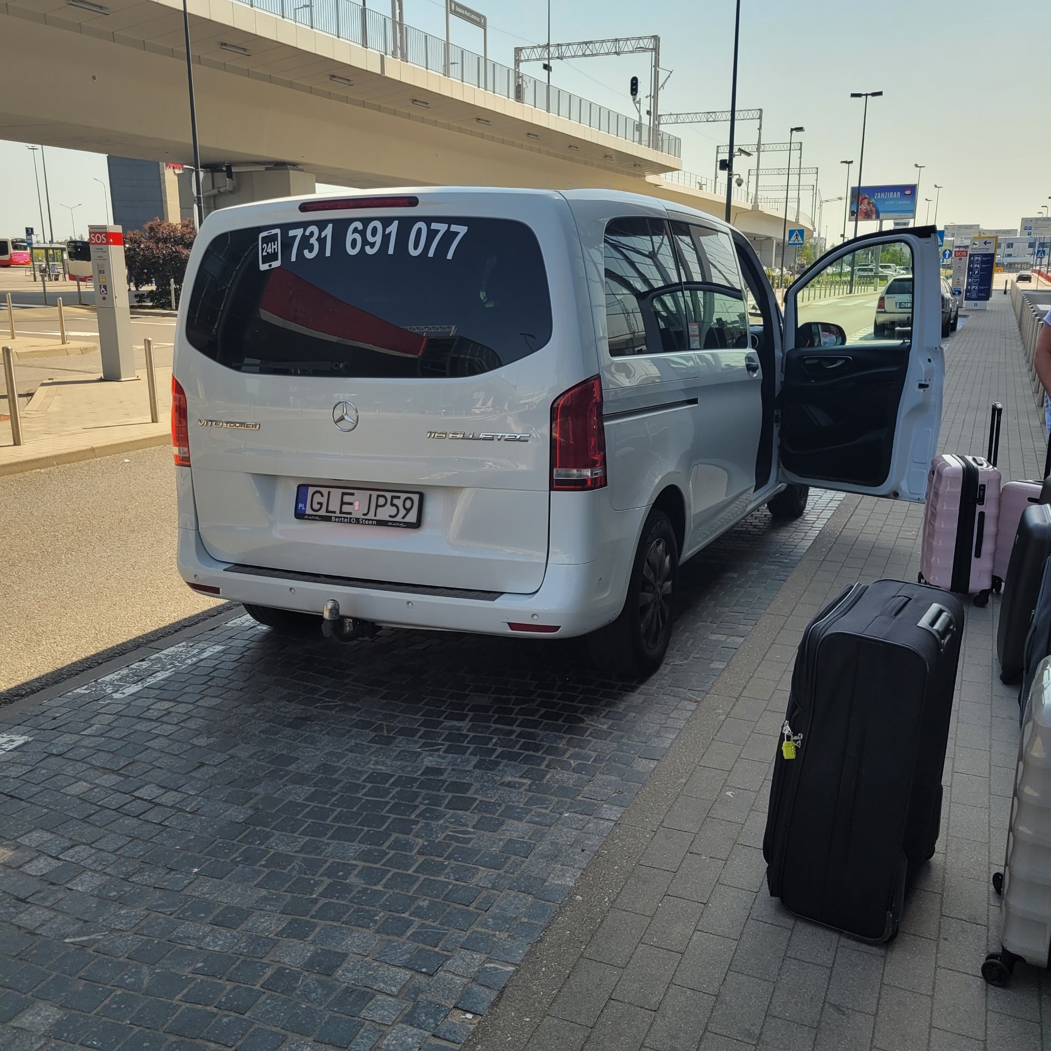 Srebrny Mercedes Vito z numerem telefonu na tylnej szybie, otwarte drzwi, bagaże obok, lotnisko w tle.