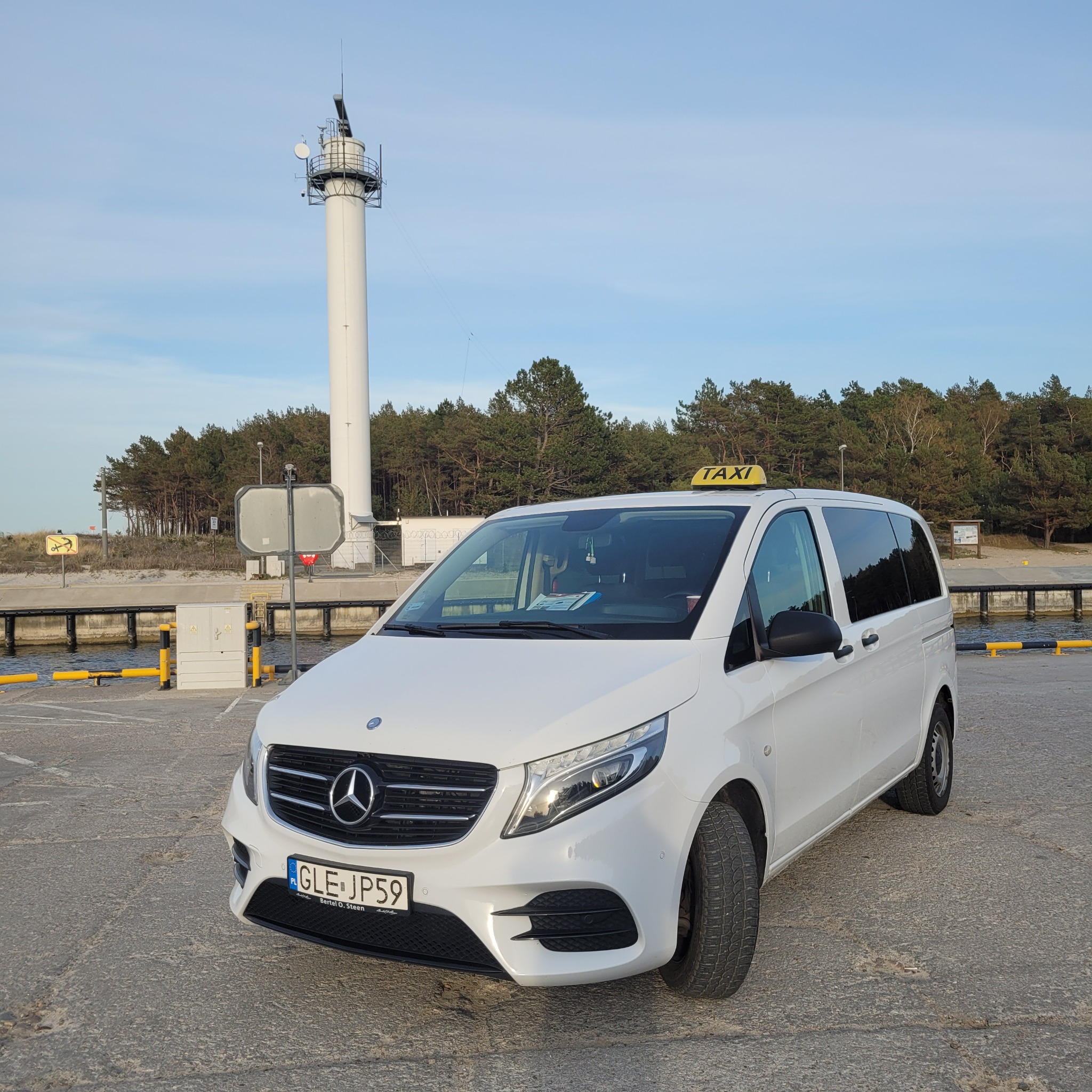Biały Mercedes Vito Taxi z żółtym oznaczeniem na dachu, zaparkowany na betonowym nabrzeżu w Łebie, w tle latarnia morska i las.
