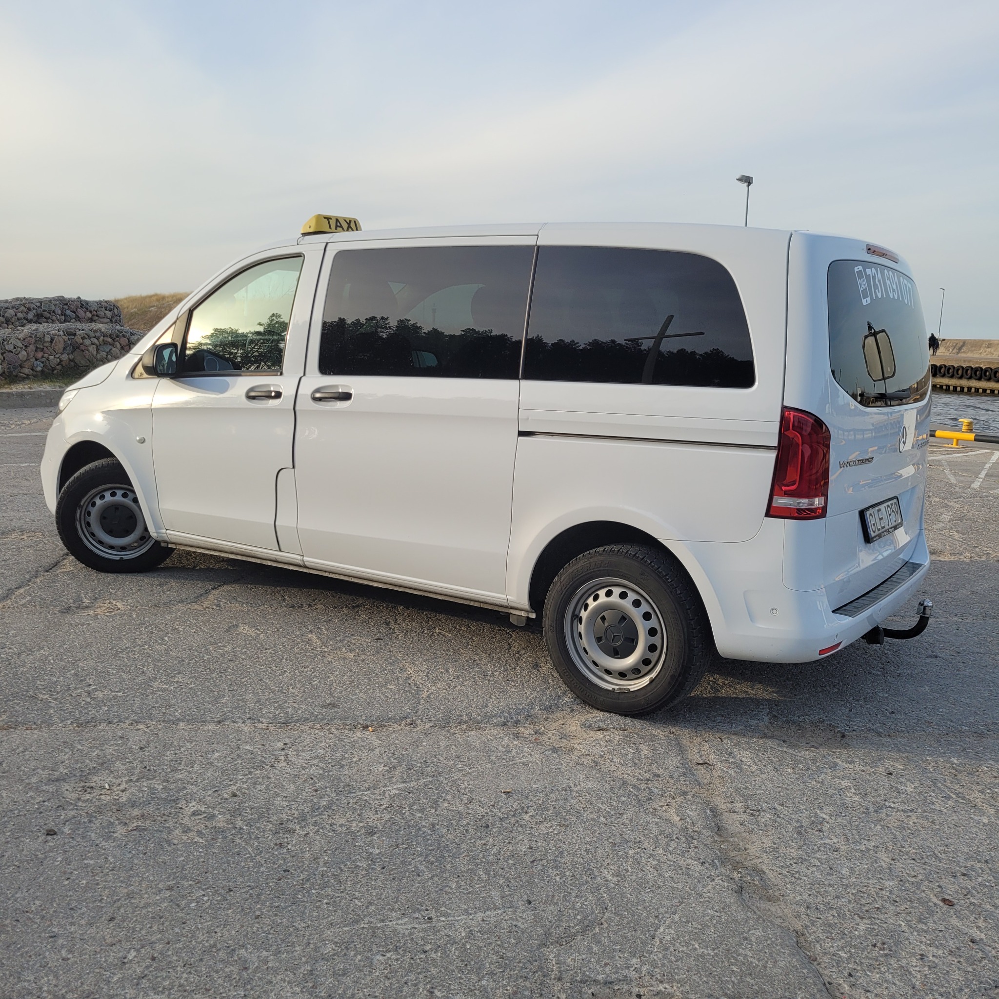 Biały samochód typu van z napisem TAXI na dachu, zaparkowany w Łebie, z widocznym numerem telefonu na tylnej szybie i z elementami nabrzeża w tle.