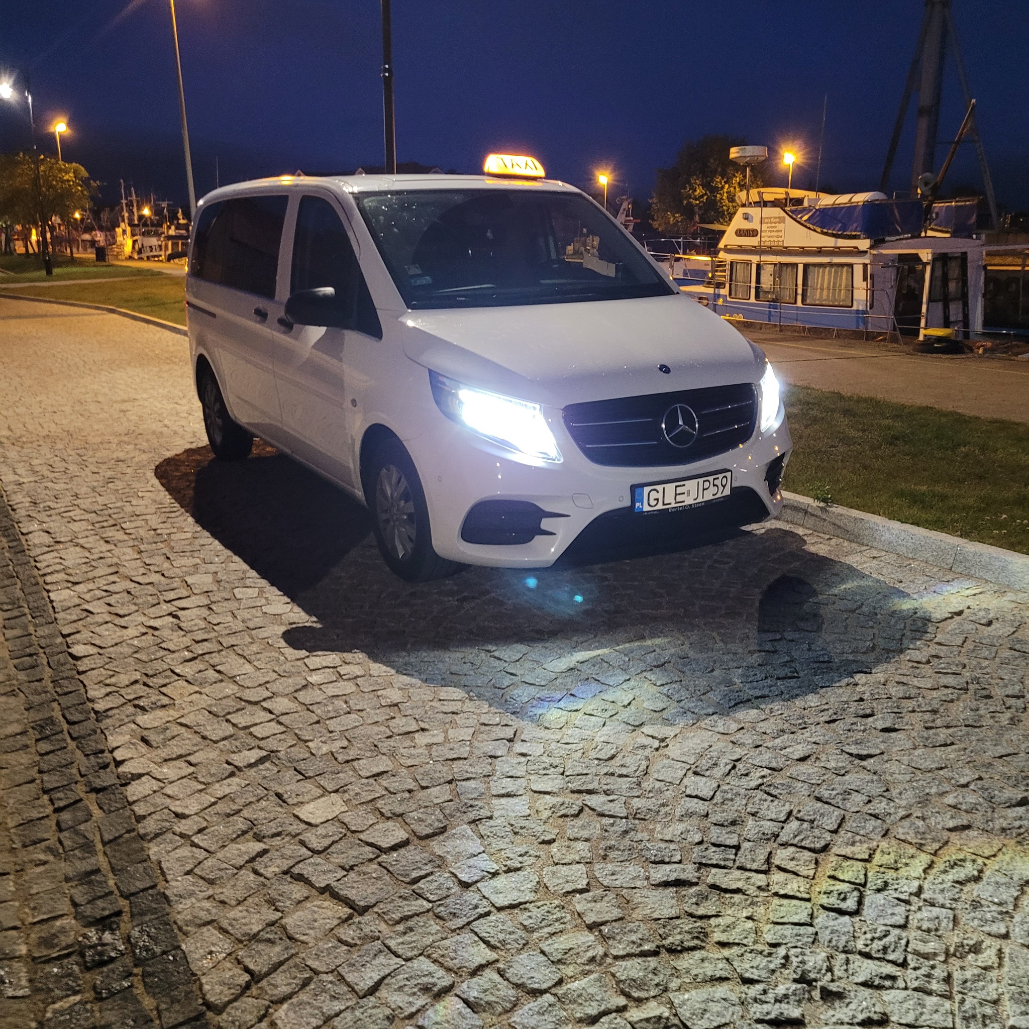 Biały Mercedes-Benz V-klasy z napisem TAXI na dachu, zaparkowany na brukowanym placu w Łebie, w tle zacumowane łodzie i wieczorne oświetlenie.