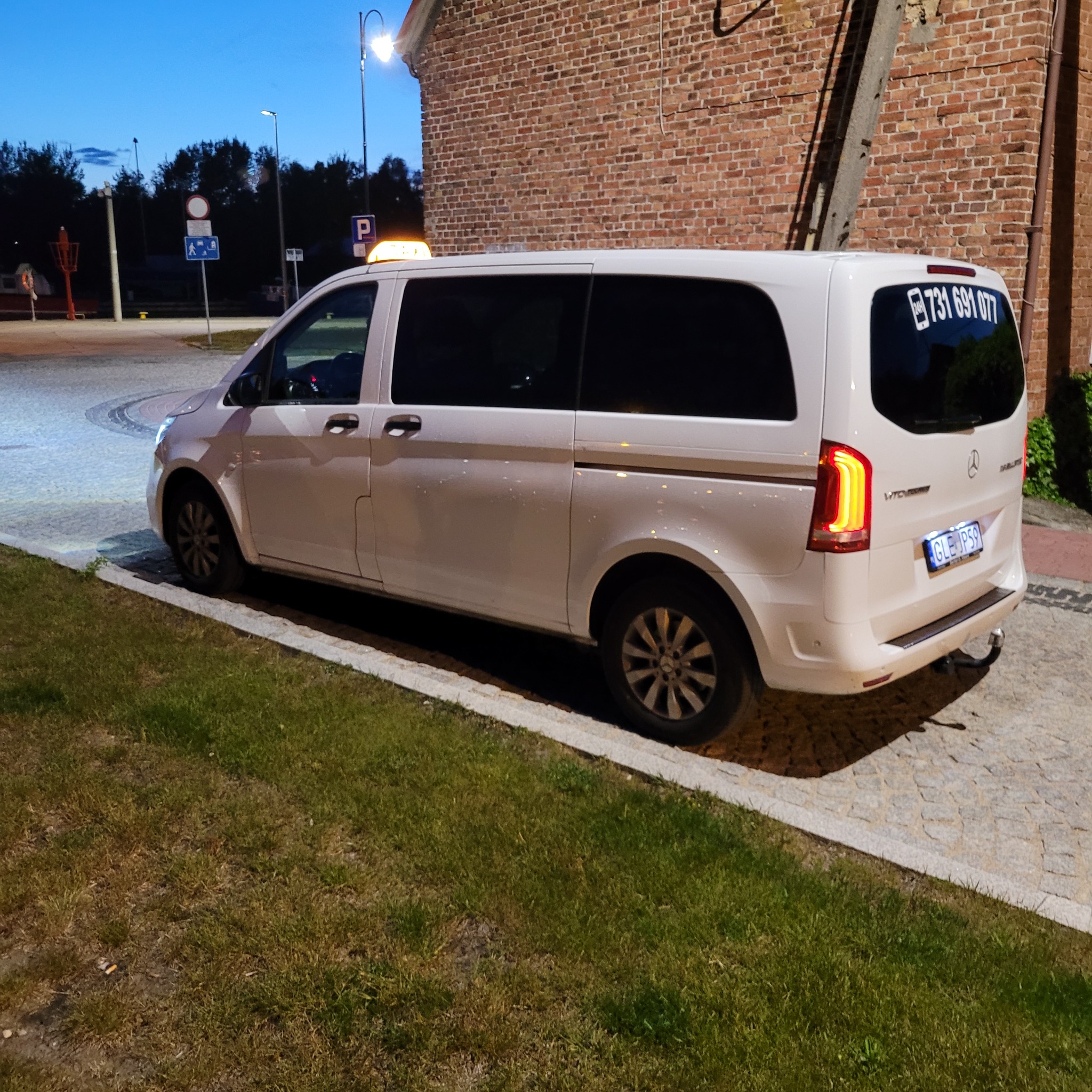 Biały van Mercedes Vito z numerem telefonu na tylnej szybie, stojący na kostce brukowej obok trawnika, z włączonymi światłami pozycyjnymi, w tle budynek z czerwonej cegły i znak taxi.