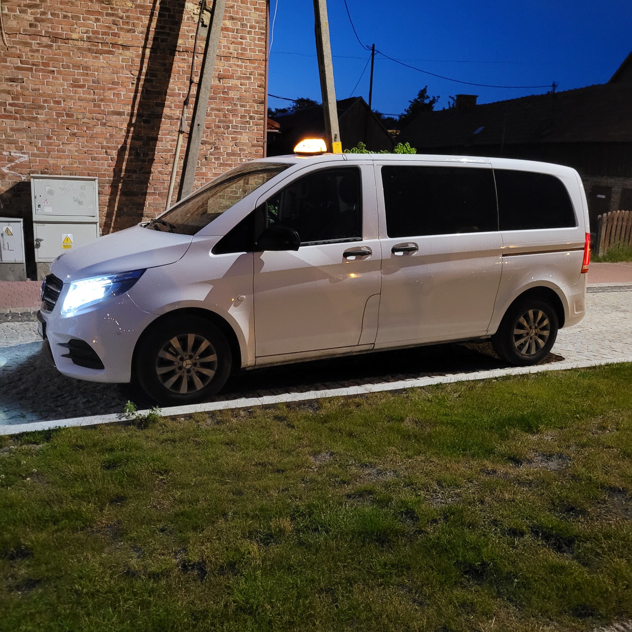Biały van z lampą Taxi na dachu, zaparkowany na chodniku z kostki brukowej przy trawniku, w tle ceglany budynek i słup oświetleniowy.