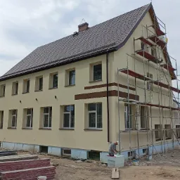 Dwupiętrowy dom z beżową elewacją i brązowym dachem podczas prac wykończeniowych; widoczne rusztowania, palety z materiałami budowlanymi i pracownik przygotowujący się do malowania elewacji...