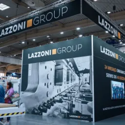 Stoisko targowe firmy Lazzoni Group z grafiką przedstawiającą detale maszyn przemysłowych, hasłami reklamowymi o gwarancji i serwisie oraz zdjęciem zespołu.