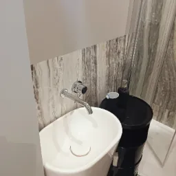 Nowoczesna łazienka z białą umywalką na postumencie, chromowaną baterią i czarnym stolikiem nocnym przypominającym beczkę, na tle marmurowej ściany i szklanej kabiny prysznicowej.