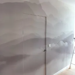 Ściana z fototapetą przedstawiającą górski krajobraz w odcieniach szarości, z ukrytymi drzwiami i motywem łodzi na dole.