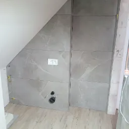 Montaż płytek imitujących marmur na skośnej ścianie w łazience, widoczne otwory na rury, puszka elektryczna i poziomujące klipsy dystansowe.