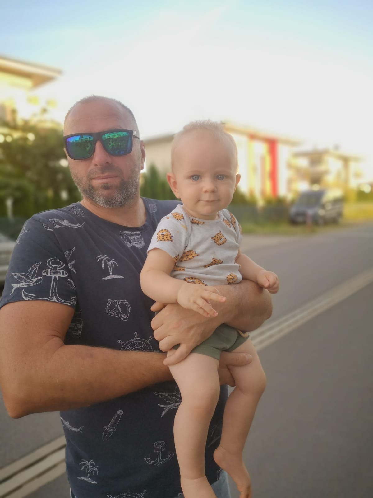 Mężczyzna w okularach przeciwsłonecznych trzymający na rękach małe dziecko na tle domu i drogi.