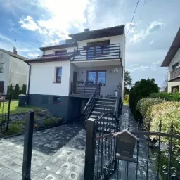 Dwupiętrowy dom z balkonami, widok zza czarnego, metalowego ogrodzenia i skrzynki na listy, na posesji ułożona szara kostka brukowa, na ścianie domu numer '6'.