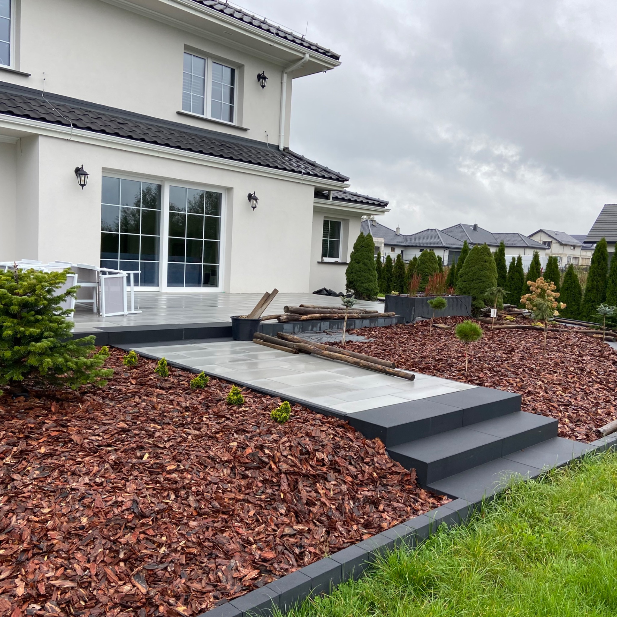 Ułożona kostka brukowa w kolorze szarym prowadząca do tarasu nowoczesnego domu, rabaty wysypane korą, na tarasie białe meble ogrodowe, widoczne drewniane elementy.