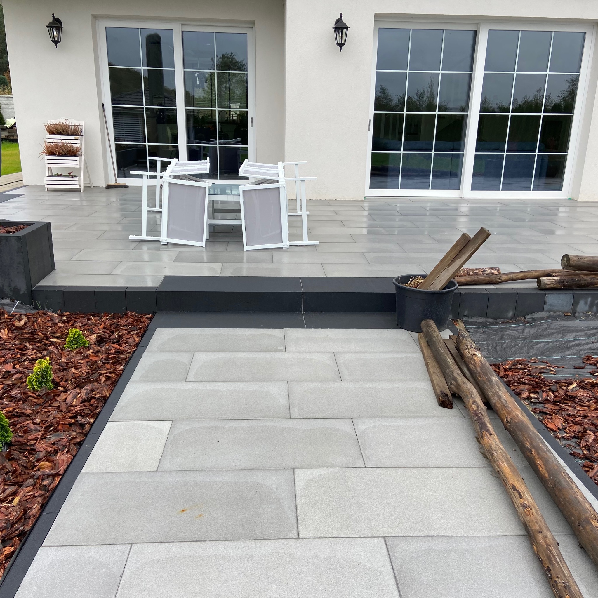 Nowoczesny taras z jasnoszarej kostki brukowej w dużym formacie, z czarnymi obrzeżami, rabata z korą i młodymi iglakami, widok na biały dom z przesuwnymi oknami, przygotowane drewno obok.