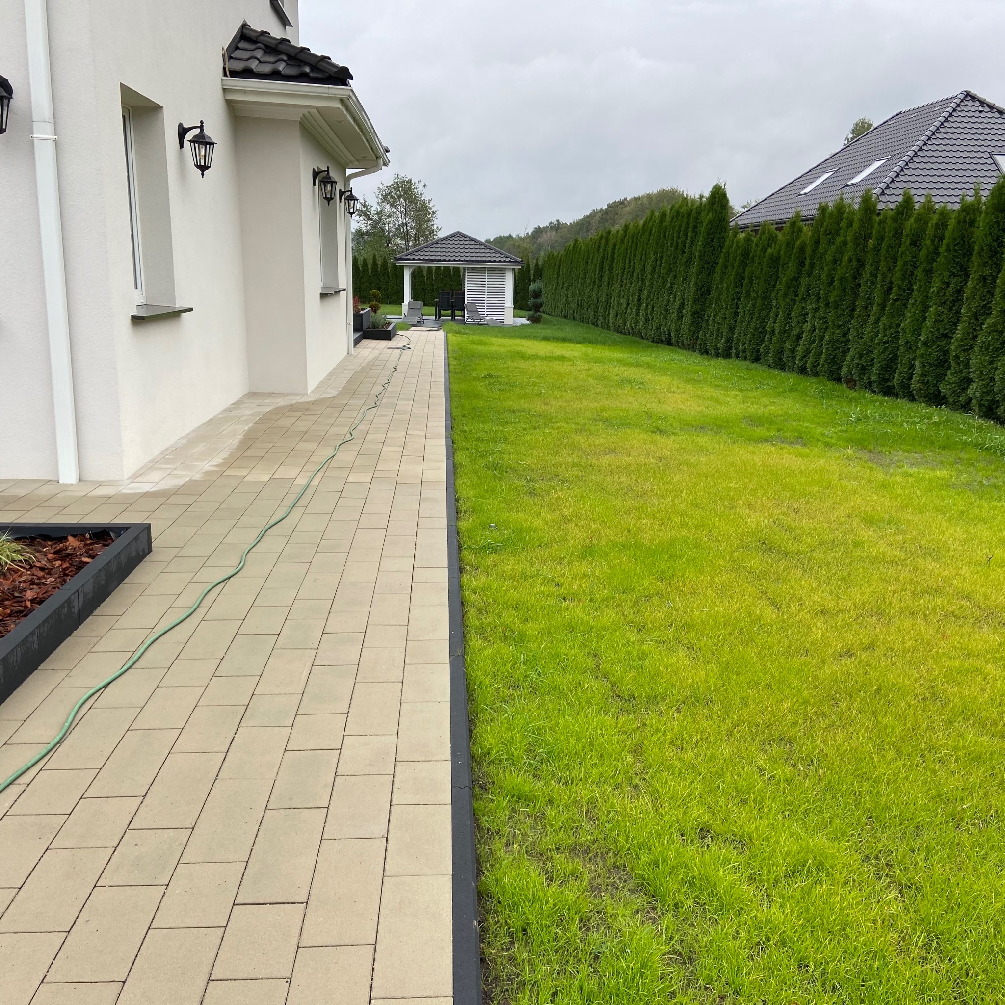 Ułożona kostka brukowa przy białym domu z czarnymi lampami, widoczny trawnik i altana w tle.