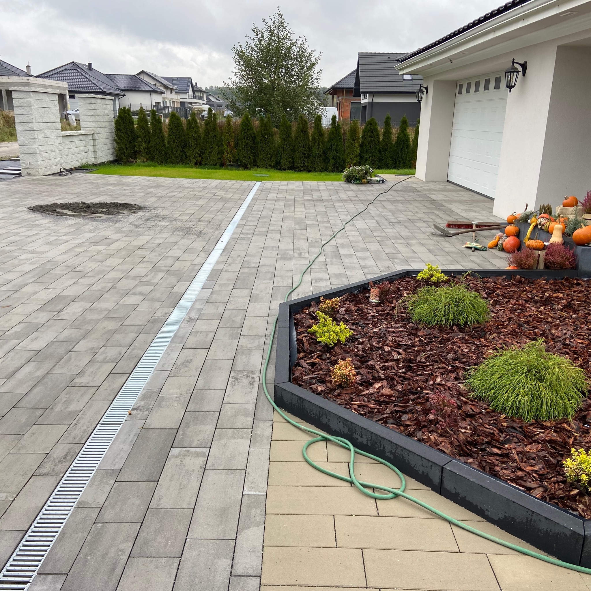 Ułożona kostka brukowa z odpływem liniowym i rabatą kwiatową z korą, dekoracje jesienne i dom w tle.