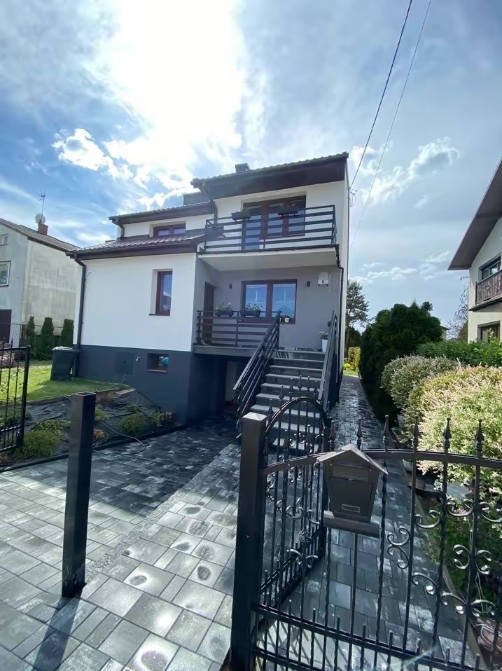 Dwupiętrowy dom z balkonami, widok zza czarnego, metalowego ogrodzenia i skrzynki na listy, na posesji ułożona szara kostka brukowa, na ścianie domu numer '6'.