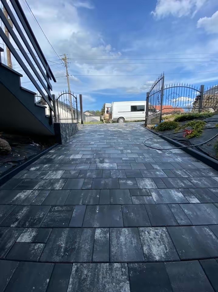 Nowo ułożona, szara kostka brukowa na podjeździe, z elementami dekoracyjnymi w postaci kutych bram i schodów z czarną balustradą, w tle biały bus i błękitne niebo.