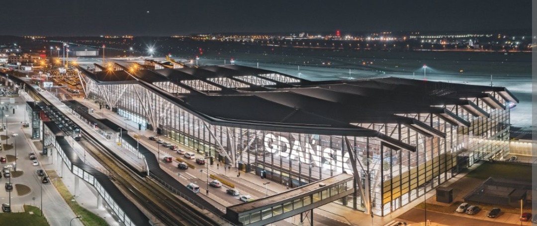 Nocny widok na oświetlony budynek lotniska w Gdańsku z widocznymi peronami kolejowymi i drogą dojazdową.