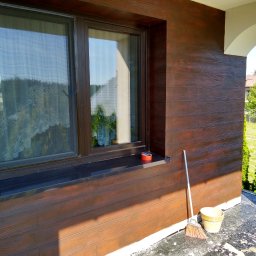 Krzysztof Remonty Bełchatów - Elewacja budynku z brązowymi panelami imitującymi drewno, widoczne okno z moskitierą, wiadro i miotła na balkonie, fragment budynku z łukiem.