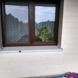 Krzysztof Remonty Bełchatów - Okna z brązowymi ramami w białej elewacji imitującej drewno, na parapecie listwa montażowa, widoczne ślady prac wykończeniowych.