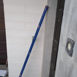 Krzysztof Remonty Bełchatów - Montaż paneli elewacyjnych imitujących drewno, z użyciem poziomicy, widoczna surowa ściana obok drzwi.