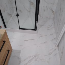 Krzysztof Remonty Bełchatów - Nowoczesna kabina prysznicowa z czarnymi profilami na tle białych płytek imitujących marmur, widoczna również drewniana szafka z czarnymi uchwytami i białe rury.