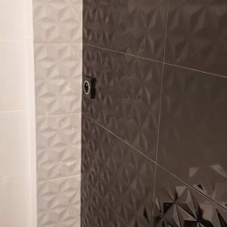 Krzysztof Remonty Bełchatów - Ściana łazienki wyłożona białymi i grafitowymi płytkami z trójwymiarowym wzorem, widoczny fragment instalacji elektrycznej.