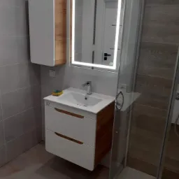 Nowoczesna łazienka z wiszącą szafką z umywalką, podświetlanym lustrem i kabiną prysznicową z przeźroczystego szkła.