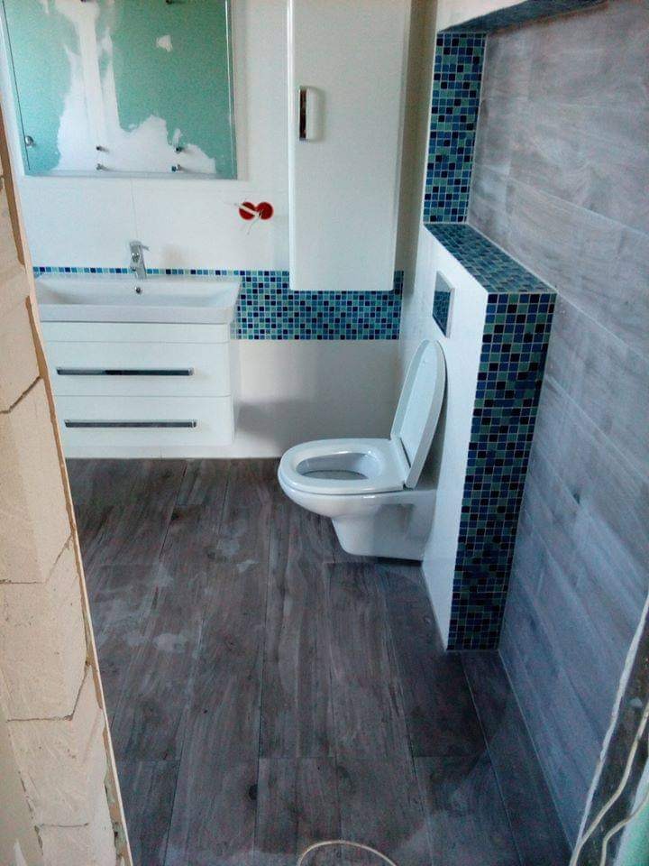 Wykończona łazienka z drewnopodobną podłogą, białą szafką z umywalką, toaletą i dekoracyjną mozaiką w odcieniach niebieskiego i granatu.