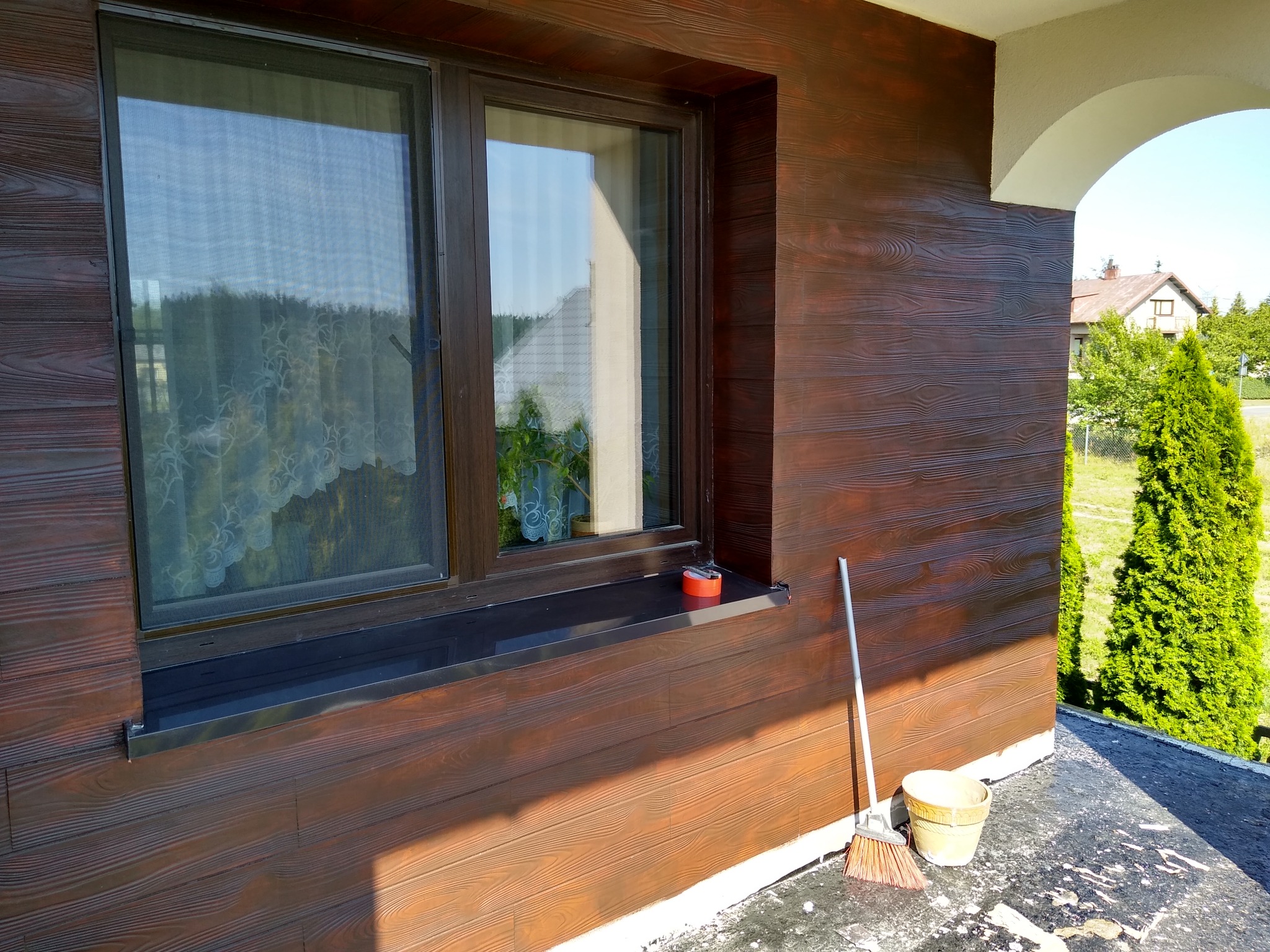 Elewacja budynku z brązowymi panelami imitującymi drewno, widoczne okno z moskitierą, wiadro i miotła na balkonie, fragment budynku z łukiem.