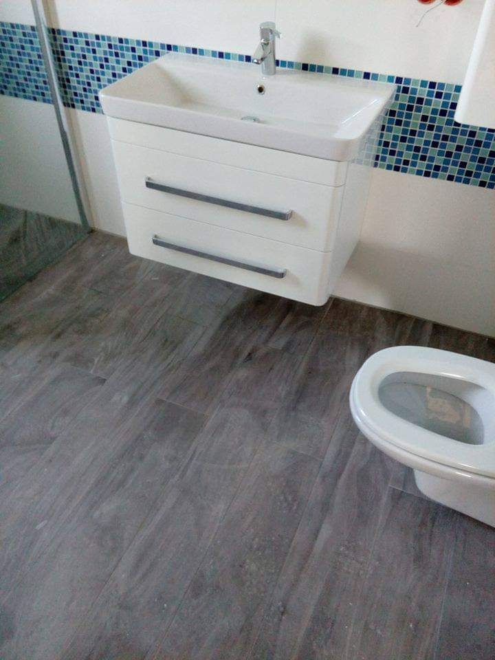 Wykończona łazienka z szarymi panelami podłogowymi imitującymi drewno, białą szafką pod umywalką z dwoma szufladami i mozaiką w odcieniach błękitu na ścianie.