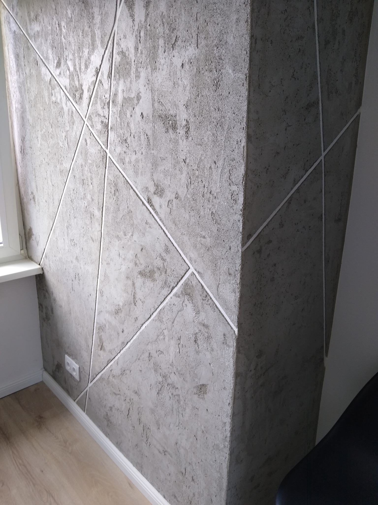 Dekoracyjna ściana w szarym odcieniu z geometrycznym wzorem z białych listew, widoczny fragment okna i podłogi z paneli.