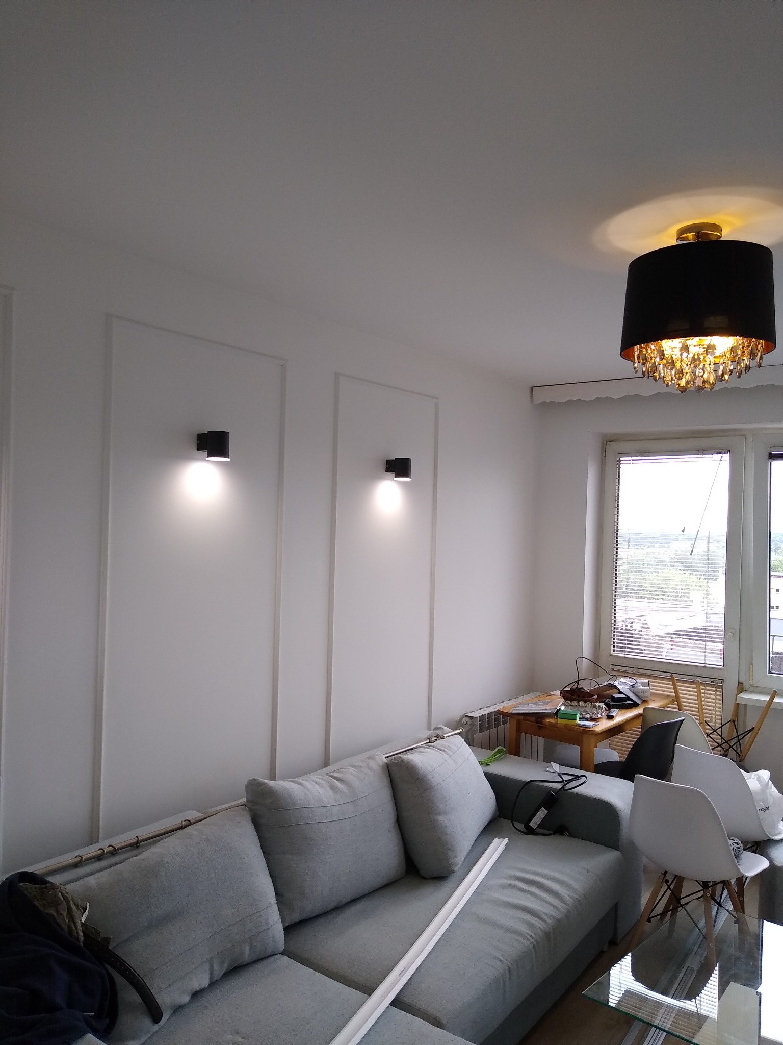 Pokój po odświeżeniu ścian z dekoracyjnymi listwami i nowymi lampami ściennymi, widoczny szary narożnik i częściowo umeblowanie.