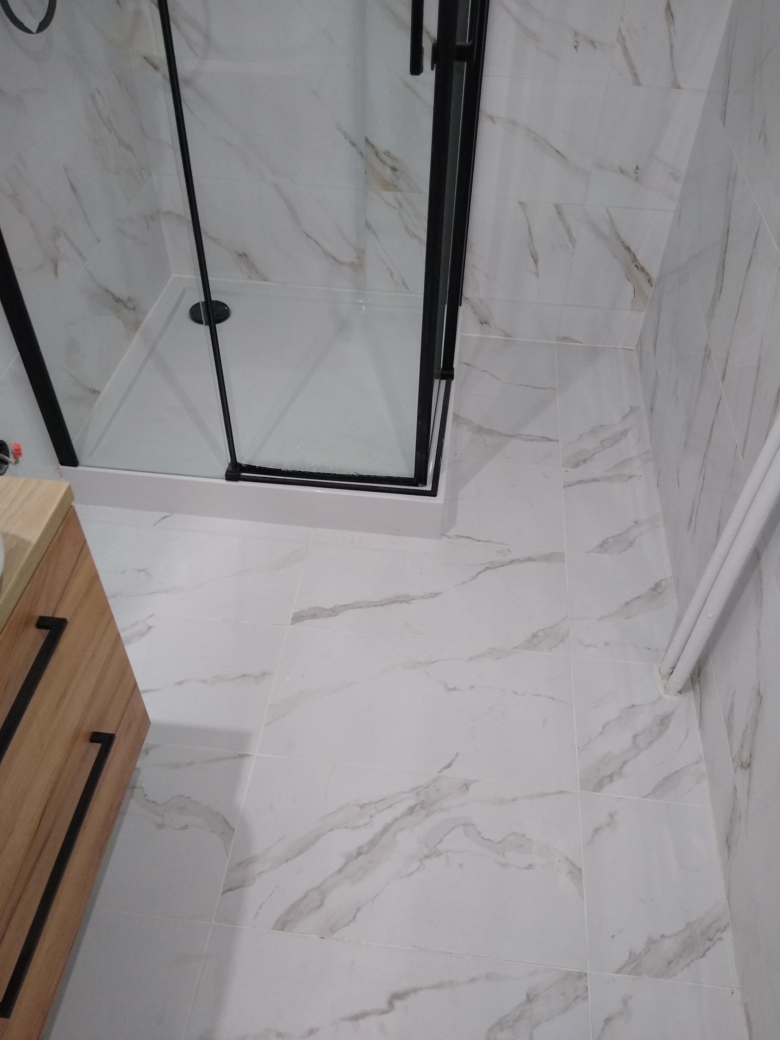 Nowoczesna kabina prysznicowa z czarnymi profilami na tle białych płytek imitujących marmur, widoczna również drewniana szafka z czarnymi uchwytami i białe rury.